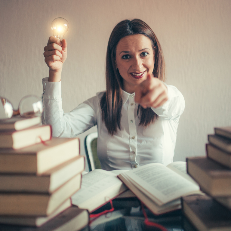 Individual seated at a desk surrounded by books, holding a light bulb, representing the power of knowledge and inspiration.